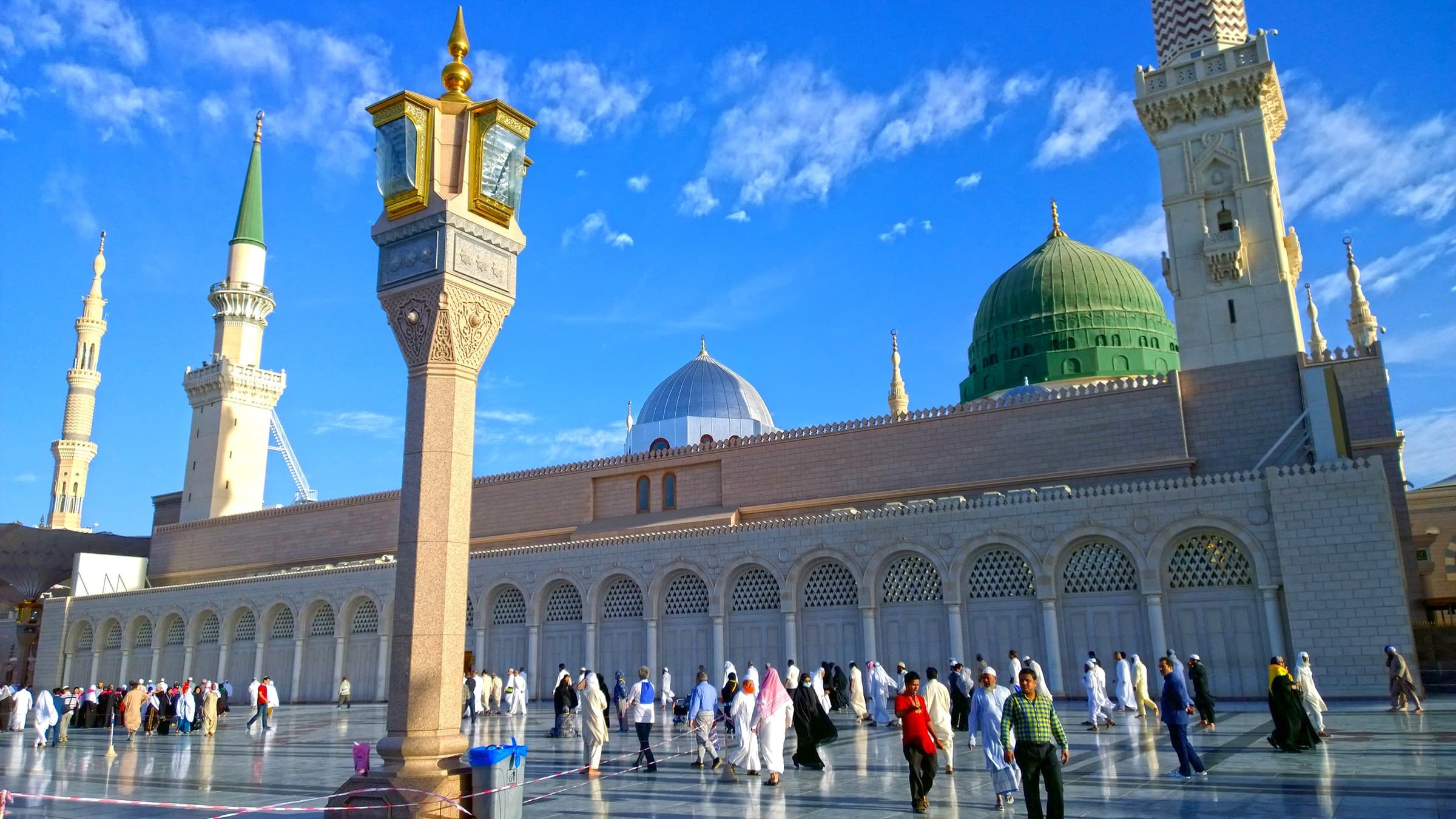 The Green Dome and minarets of Masjid an-Nabawi in Madinah under a blue sky, with visitors in the courtyard